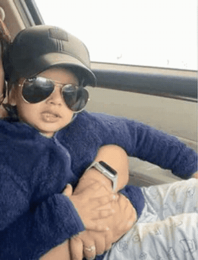 A Woman Is Holding A Baby In Her Arms In A Car . The Baby Is Wearing Sunglasses And A Baseball Cap . GIF