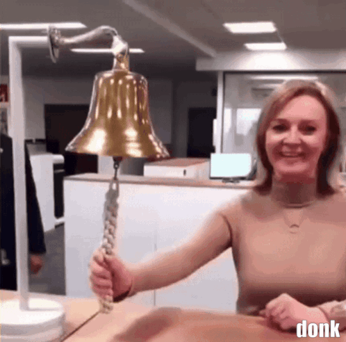 A Woman Is Holding A Bell In Front Of A Sign That Says ' Donk ' On It GIF