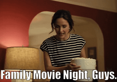 A Woman Is Holding A Cake And A Bucket Of Popcorn And The Caption Says Family Movie Night Guys GIF