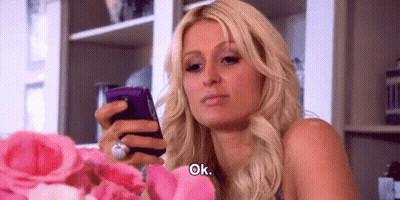 A Woman Is Holding A Cell Phone In Her Hand While Sitting In Front Of A Bouquet Of Pink Roses . GIF