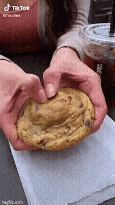 A Woman Is Holding A Cookie In Her Hands With A Cup Of Coffee In The Background . GIF