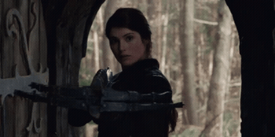 A Woman Is Holding A Crossbow In Front Of A Door In A Forest . GIF