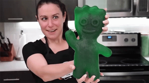 A Woman Is Holding A Giant Green Gummy Bear In Front Of A Stove GIF