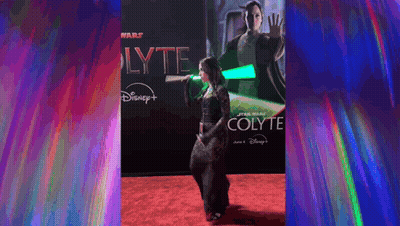 a woman is holding a green lightsaber on a red carpet in front of a poster for star wars colyte .