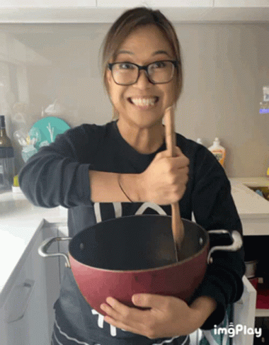 A Woman Is Holding A Pot With A Wooden Spoon In It And Smiling GIF