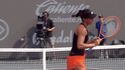 A Woman Is Holding A Tennis Racquet On A Court In Front Of A Wall That Says Caliente GIF