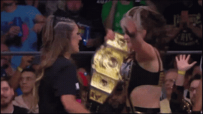 A Woman Is Holding A Wrestling Championship While Another Woman Watches GIF