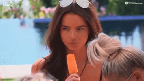 A Woman Is Holding An Orange Popsicle In Her Hand While Wearing Sunglasses . GIF