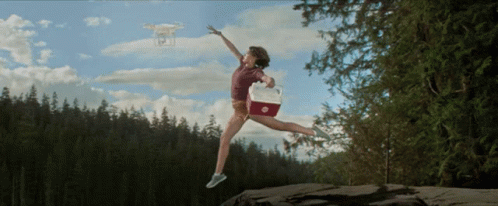 A Woman Is Jumping In The Air With A Cooler And A Drone In The Background GIF