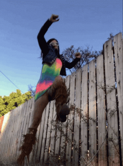 A Woman Is Jumping Over A Wooden Fence Wearing A Colorful Dress GIF