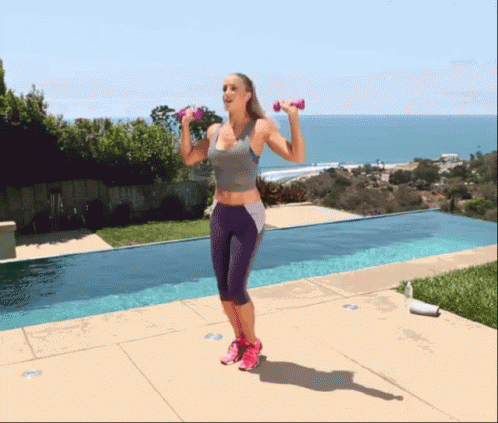 A Woman Is Jumping With Dumbbells In Front Of A Pool . GIF