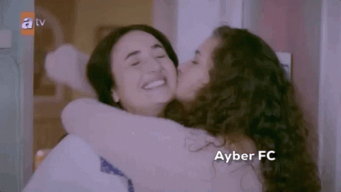 A Woman Is Kissing Another Woman On The Cheek While Hugging Each Other . GIF
