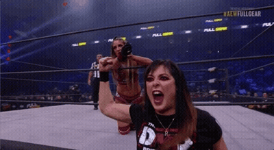 A Woman Is Kneeling Down In A Wrestling Ring While Another Woman Holds Her Fist Up In The Air . GIF