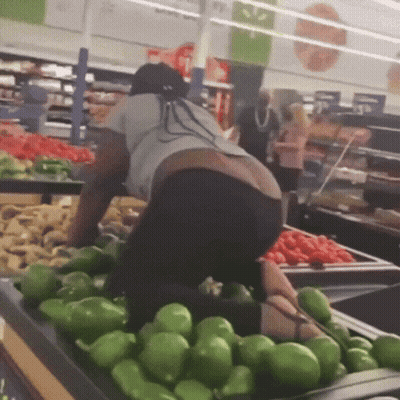 A Woman Is Kneeling Down In Front Of A Bunch Of Green Peppers In A Grocery Store GIF