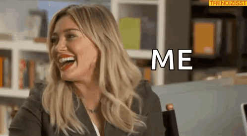 A Woman Is Laughing And Saying `` Me '' While Sitting In Front Of A Bookshelf . GIF