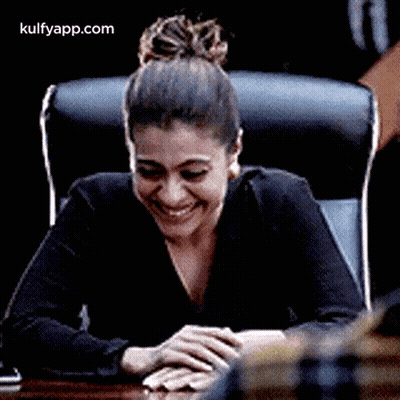 A Woman Is Laughing While Sitting At A Table With Her Hands On Her Lap . GIF