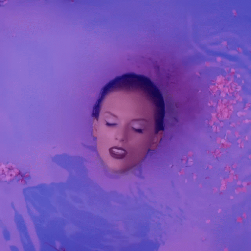A Woman Is Laying In A Bathtub With Flowers Falling Around Her GIF