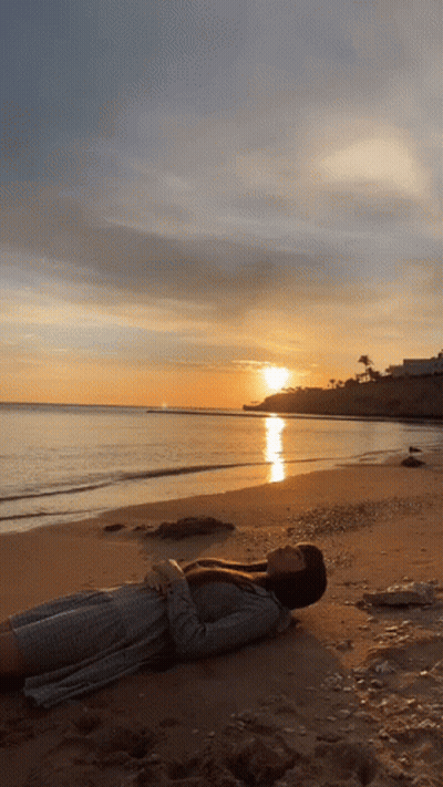 A Woman Is Laying On A Beach With The Sun Setting In The Background GIF