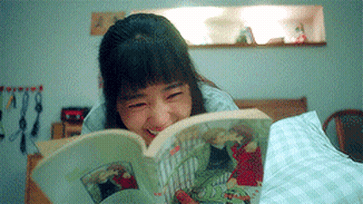 A Woman Is Laying On A Bed Reading A Book And Smiling . GIF