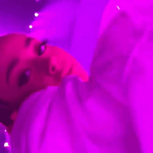 A Woman Is Laying On A Bed Under A Purple Light And Looking At The Camera . GIF