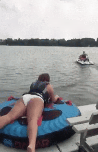 A Woman Is Laying On A Tube In The Water With A Jet Ski Behind Her . GIF