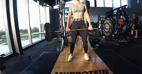 A Woman Is Lifting A Barbell In A Gym That Has The Word Protein On The Floor GIF