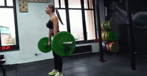 A Woman Is Lifting A Barbell In A Gym With A Digital Clock In The Background . GIF