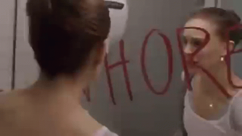 A Woman Is Looking At Her Reflection In A Mirror And The Word Whore Is Written On The Mirror . GIF