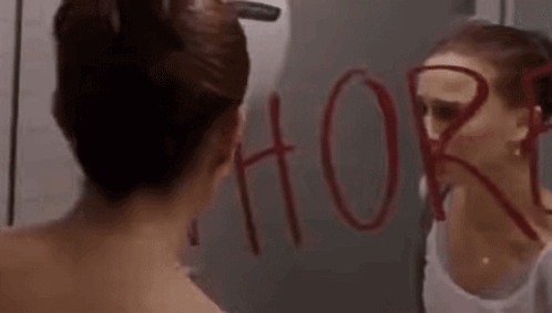 A Woman Is Looking At Herself In A Bathroom Mirror With The Word Whore Written On It . GIF