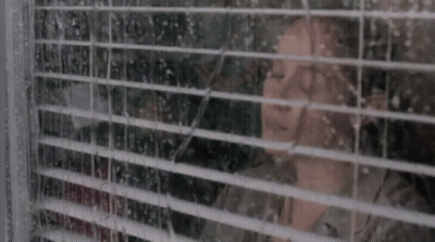 A Woman Is Looking Out A Window With Blinds On It . GIF