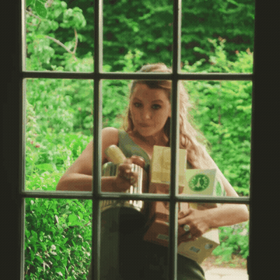 A Woman Is Looking Out Of A Window Holding A Box With A Green Circle On It GIF