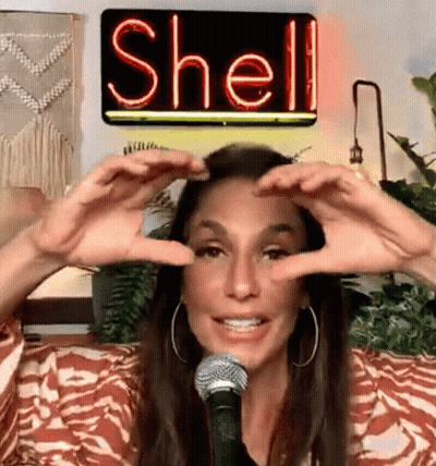 A Woman Is Making A Heart Shape With Her Hands In Front Of A Neon Sign That Says Shell On It . GIF