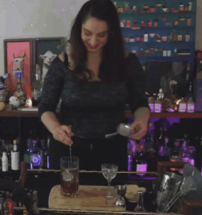 A Woman Is Pouring A Drink Into A Glass In Front Of A Shelf Of Bottles GIF
