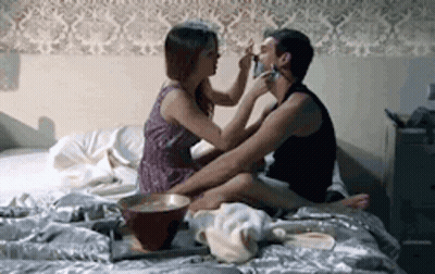 A Woman Is Putting Makeup On A Man 'S Face While They Are Sitting On A Bed . GIF