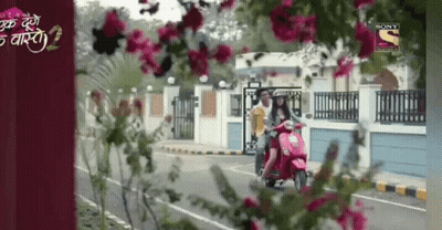 A Woman Is Riding A Scooter Down A Street With Flowers In The Foreground And A Sony Channel Logo In The Background GIF
