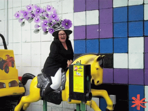 A Woman Is Riding A Yellow Horse In Front Of A Sign That Says ' Kids Play ' On It GIF