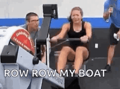A Woman Is Rowing A Boat In A Gym With A Man Standing Behind Her . GIF
