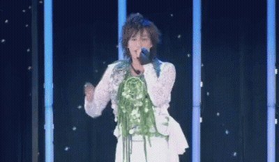 A Woman Is Singing Into A Microphone On A Stage With A Bouquet Of Flowers On Her Neck . GIF