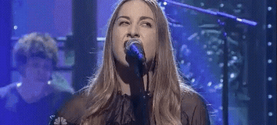 A Woman Is Singing Into A Microphone On A Stage With A Man Playing A Piano In The Background . GIF