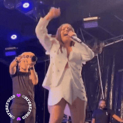 A Woman Is Singing Into A Microphone On Stage While A Man Takes A Picture Of Her . GIF