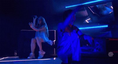 A Woman Is Singing Into A Microphone While A Man Is Kneeling Down Behind Her GIF
