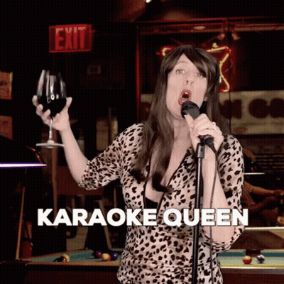 A Woman Is Singing Into A Microphone While Holding A Glass Of Wine And The Words Karaoke Queen Are Below Her GIF