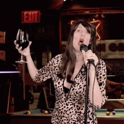 A Woman Is Singing Into A Microphone While Holding A Glass Of Wine In Front Of An Exit Sign GIF