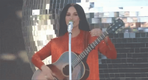 A Woman Is Singing Into A Microphone While Holding A Guitar GIF