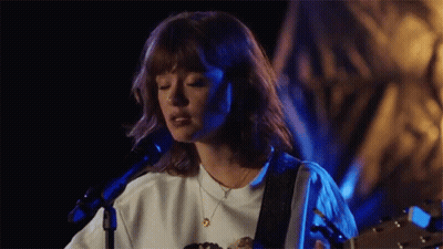 A Woman Is Singing Into A Microphone While Holding A Guitar . GIF