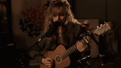 A Woman Is Singing Into A Microphone While Playing A Guitar GIF