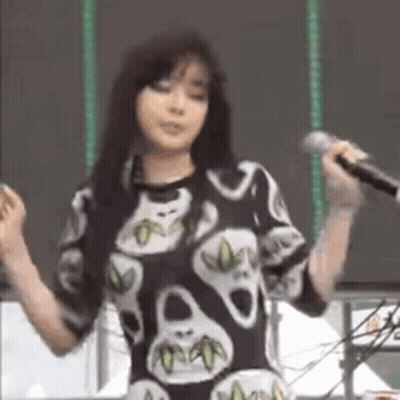 A Woman Is Singing Into A Microphone While Wearing A Skull Print Shirt . GIF