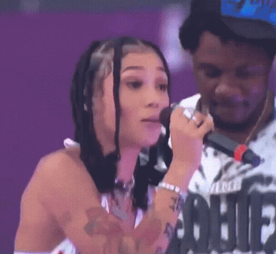 A Woman Is Singing Into A Microphone With A Man Standing Behind Her . GIF