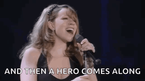 A Woman Is Singing Into A Microphone With The Words `` And Then A Hero Comes Along '' Behind Her . GIF