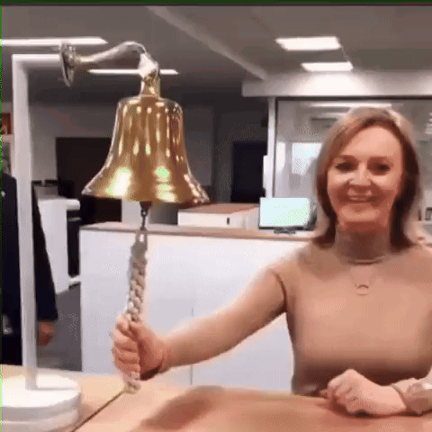 A Woman Is Sitting At A Desk Holding A Large Bell . GIF